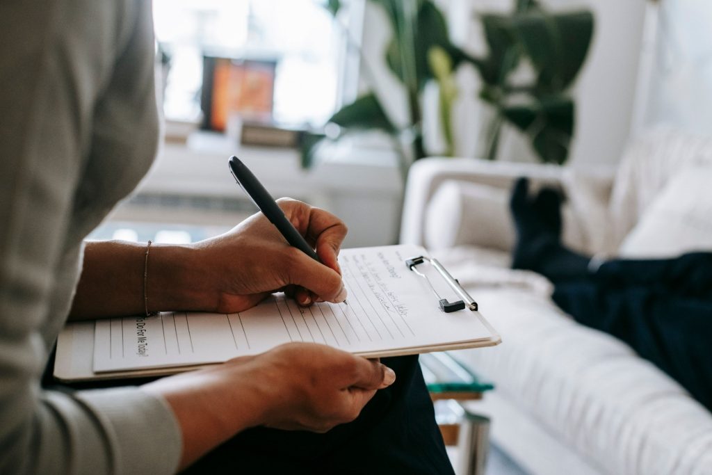 Therapist writing notes on a clipboard while a client relaxes on a couch nearby